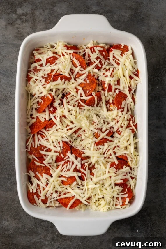 A final layer of shredded cheese is added to pizza casserole in a baking dish.