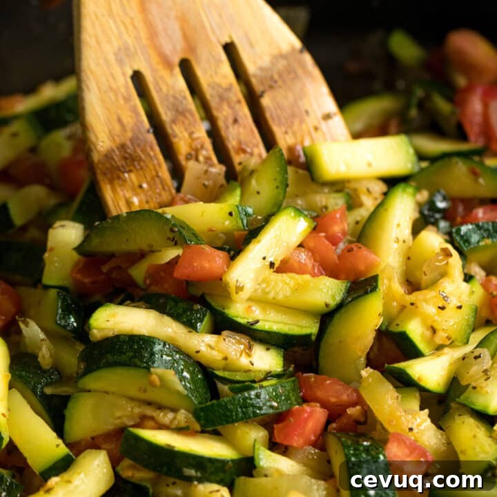A wooden spoon stirring a pan of zucchini and tomatoes.