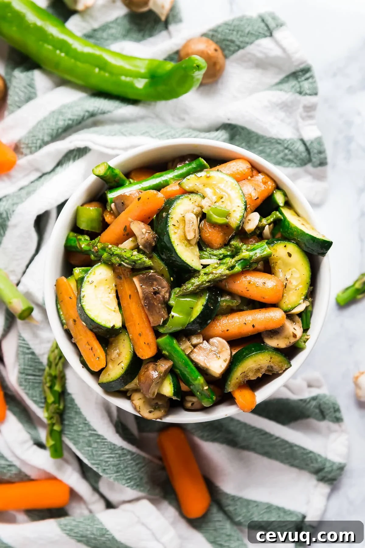 An overhead image of a bowl of sautéed veggies on top of a green and white striped towel with raw veggies around it.