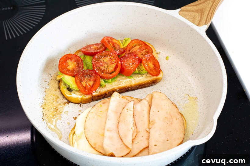 Two slices of bread in a pan, one with avocado and tomato and the other with cheese and turkey.
