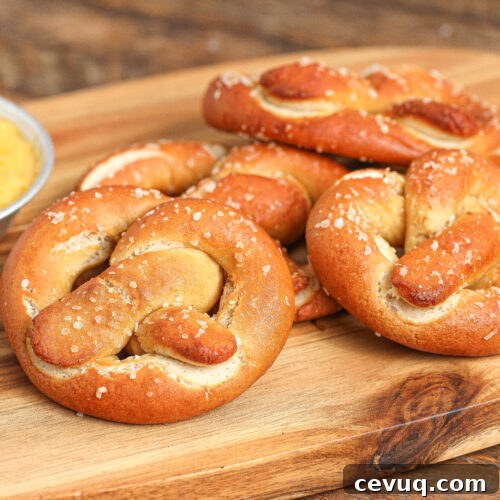 Four golden-brown soft pretzels sprinkled with coarse salt rest on a wooden board, with a small dish of yellow dipping sauce nearby.
