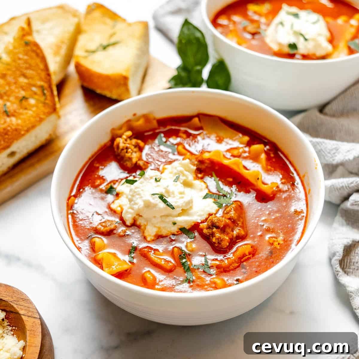 A bowl of lasagna soup topped with ricotta cheese and herbs, with slices of garlic bread and another bowl of soup in the background.
