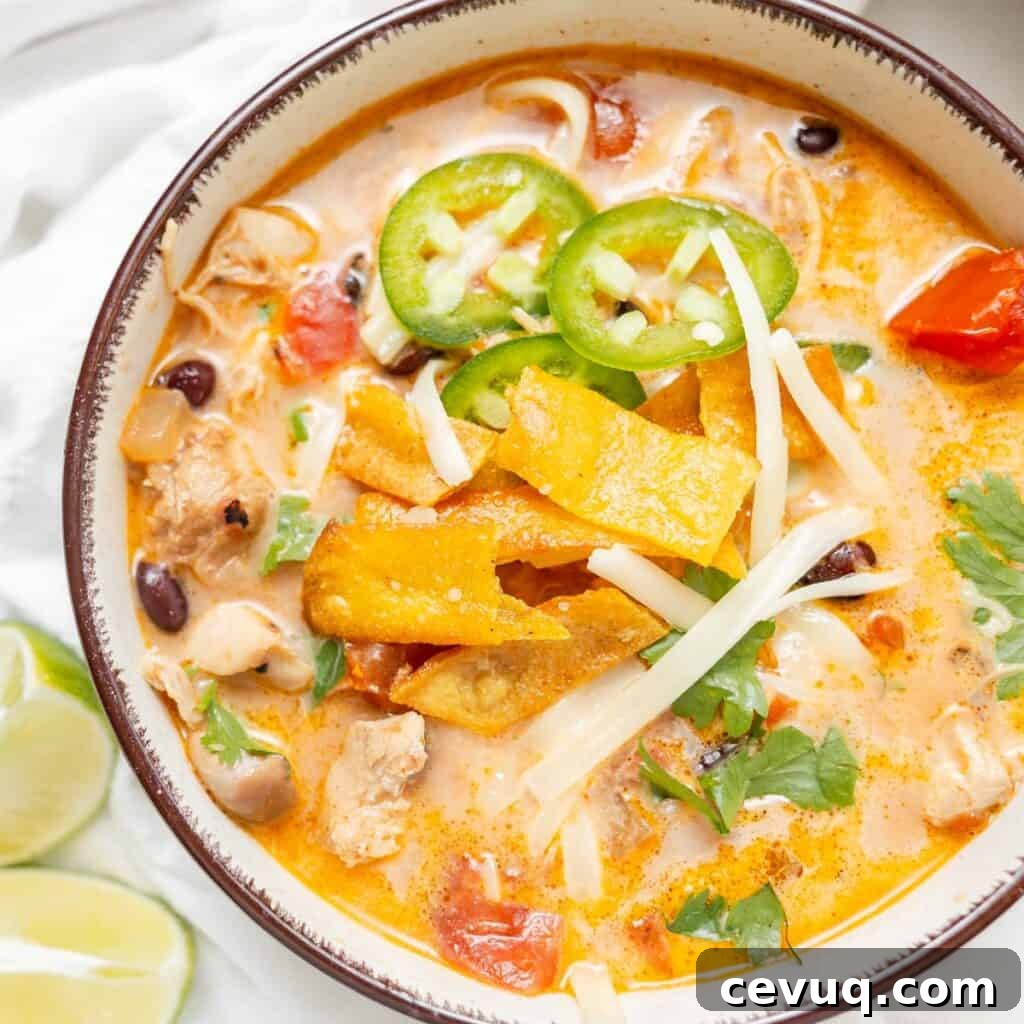 A bowl of creamy chicken tortilla soup topped with tortilla strips, sliced jalapeños, shredded cheese, and cilantro, with lime wedges on the side.