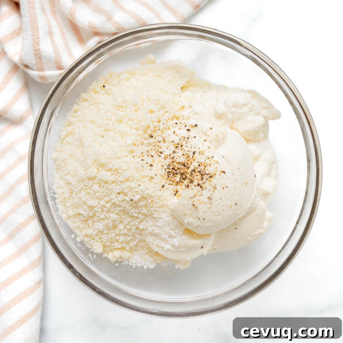 A glass bowl containing grated parmesan cheese, mayonnaise, and ground black pepper sits on a white surface next to a striped cloth.