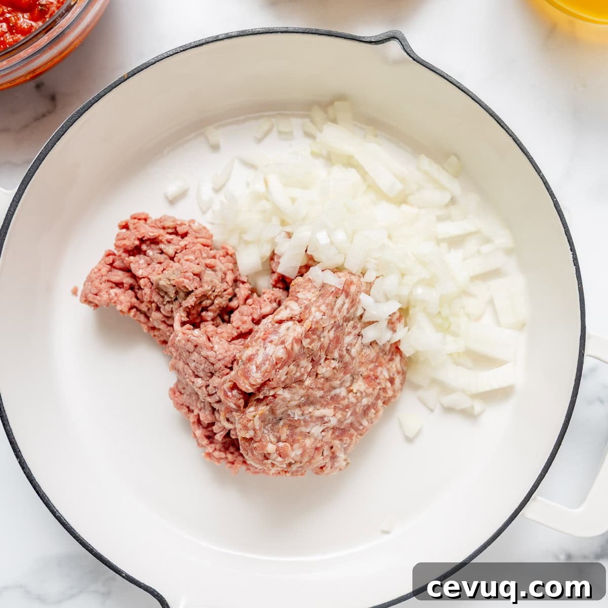 Raw ground beef and chopped onions placed in a white skillet, ready to be cooked.