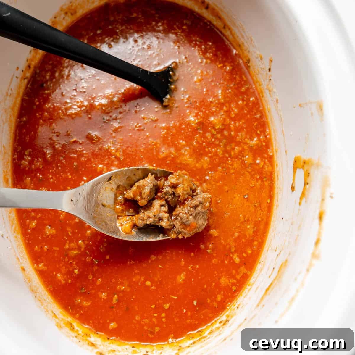 A metal spoon with cooked ground meat rests above a red tomato-based sauce in a white slow cooker, with a black utensil partially visible in the background.
