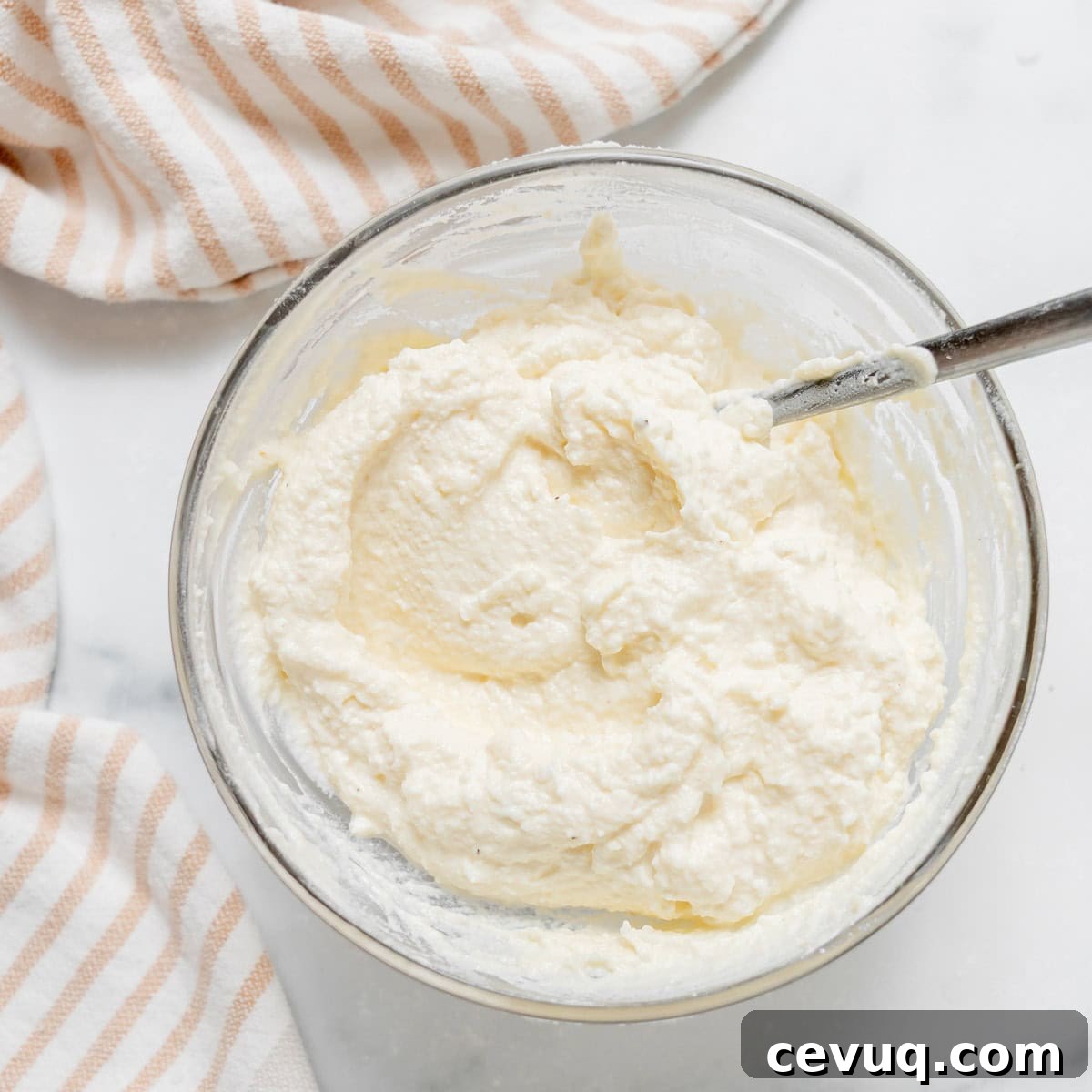 A glass bowl filled with a creamy ricotta mixture and a metal spoon, placed on a white surface next to a beige and white striped cloth.