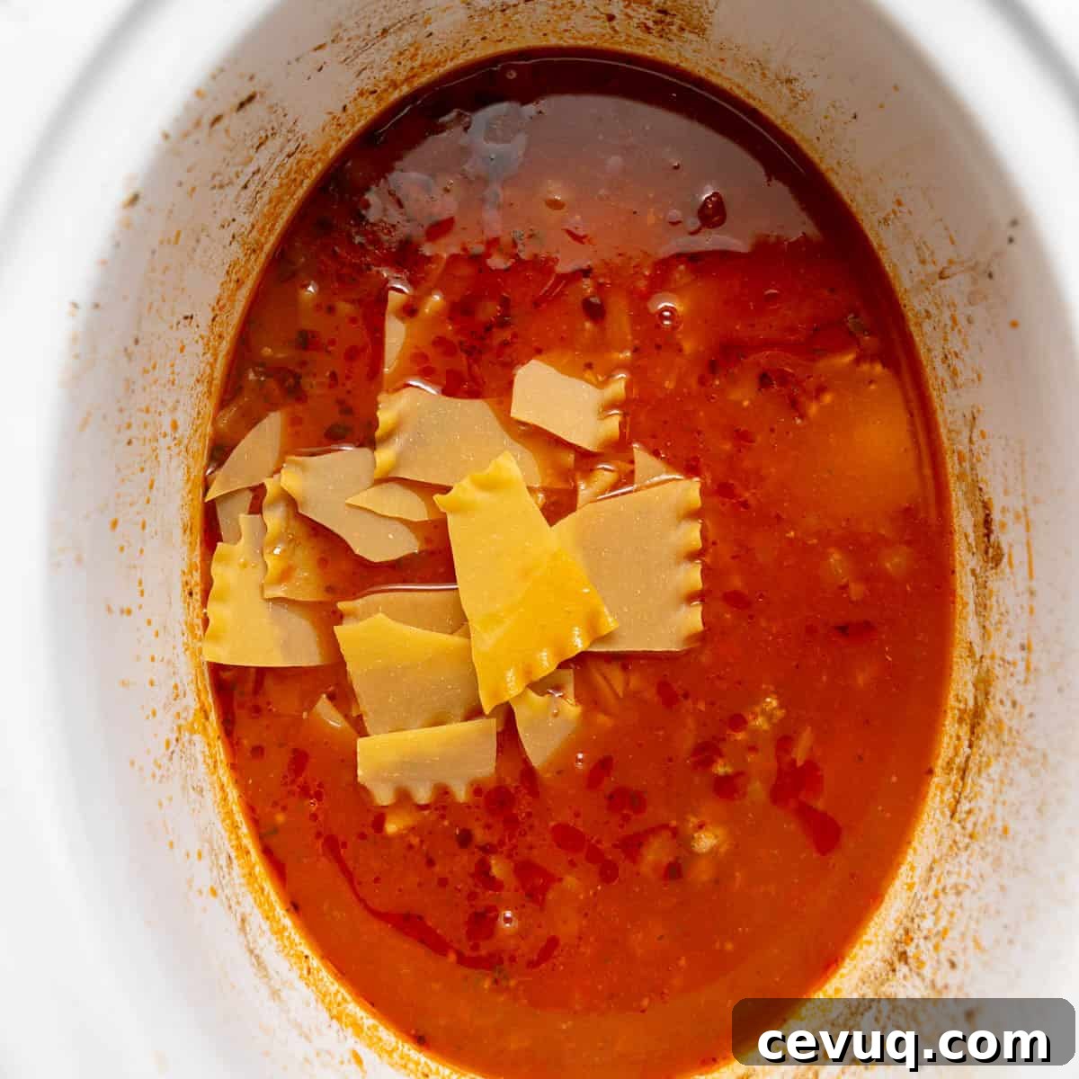 A white slow cooker filled with red broth, ground meat, and broken pieces of uncooked lasagna noodles.