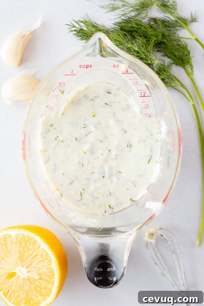 Top down shot of Greek yogurt marinade in a plastic measuring cup on top of a white background with fresh dill, garlic, and half a lemon surrounding it.