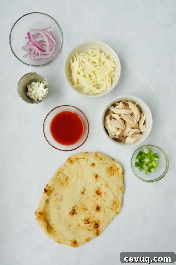 Ingredients for buffalo chicken flatbread displayed on a white marble counter including flatbread, cooked chicken, green onions, red onions, mozzarella cheese, buffalo sauce, and feta cheese