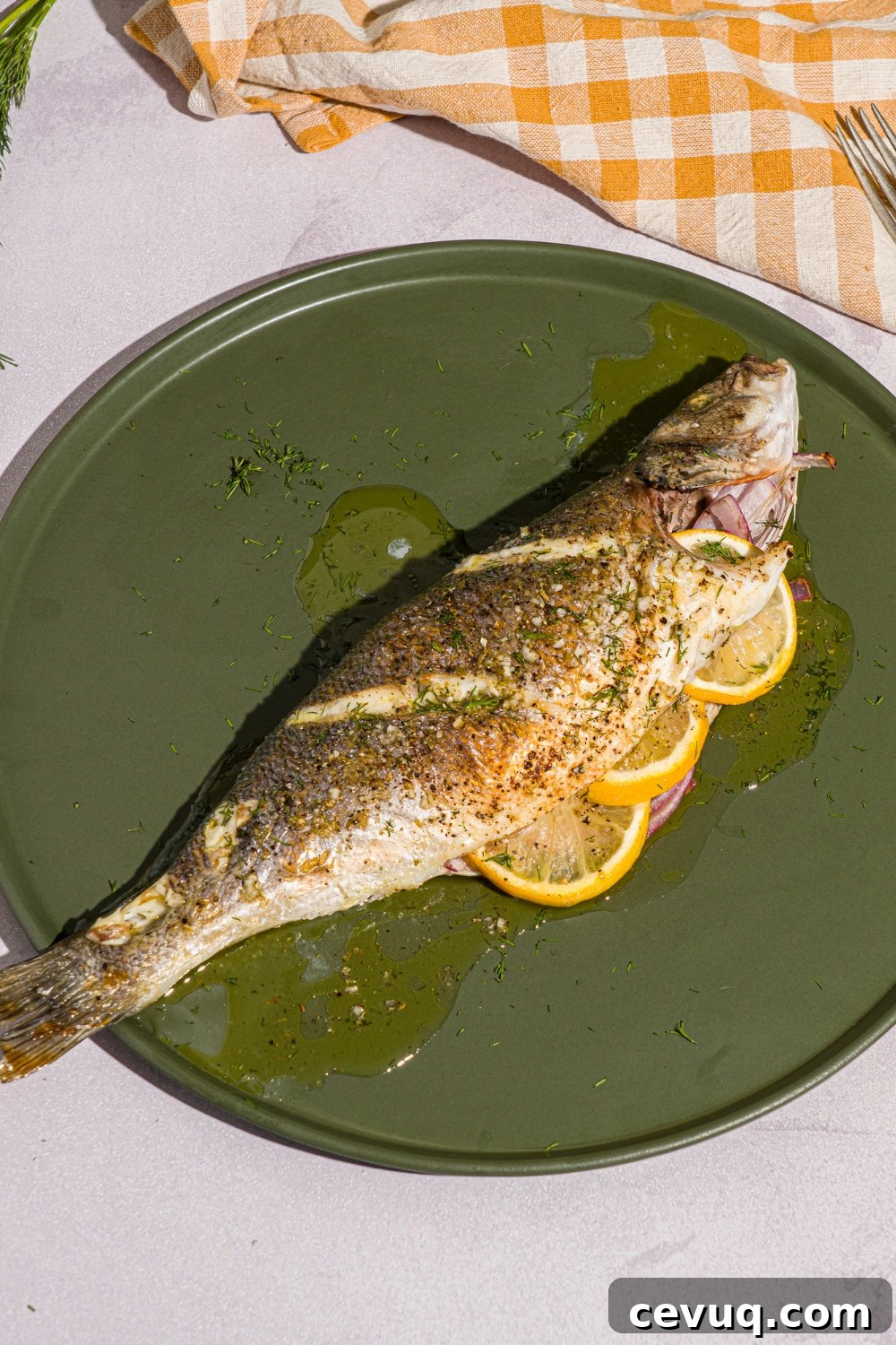 A green plate with greek whole roasted branzino stuffed with sliced lemon. The plate is on a white counter with a yellow checkered napkin.