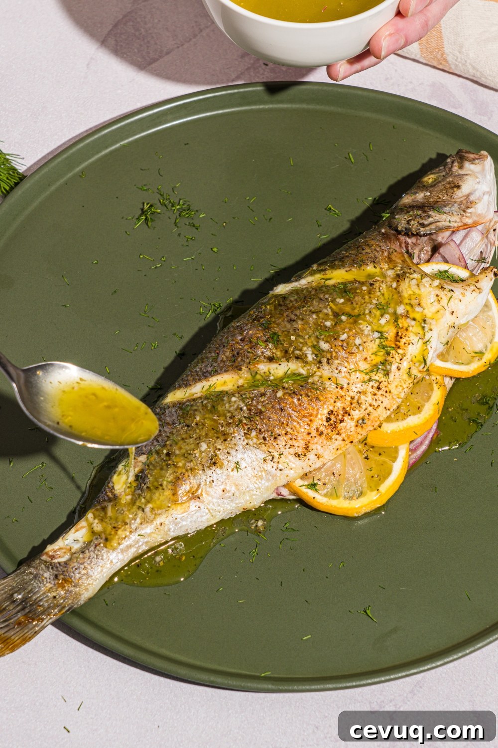 A green plate with greek whole roasted branzino stuffed with sliced lemon. A spoon is drizzling ladolemono dressing over the branzino. The plate is on a white counter with a yellow checkered napkin.