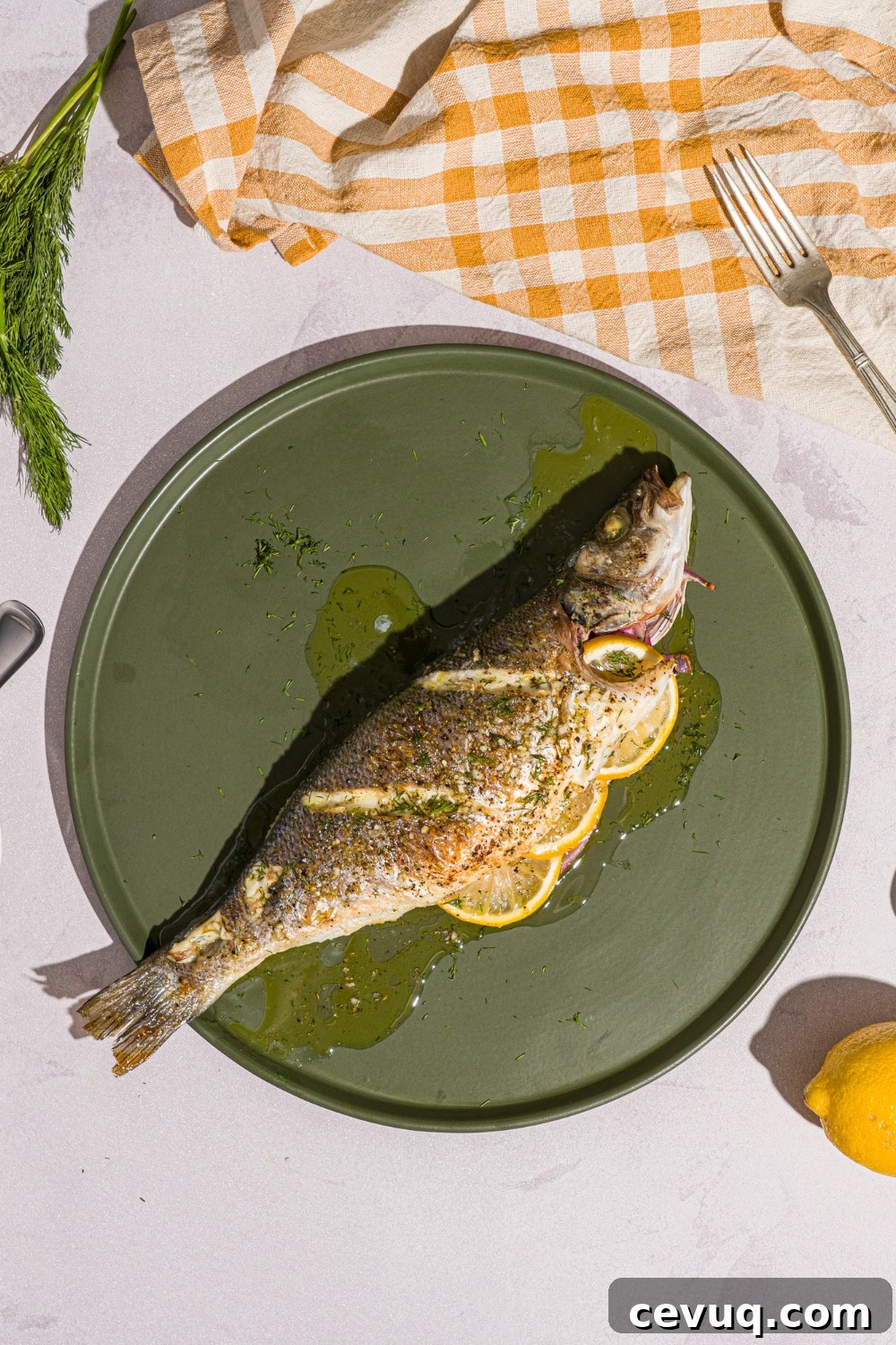 A green plate with greek whole roasted branzino stuffed with sliced lemon and drizzled with ladolemono dressing. The plate is on a white counter with a yellow checkered napkin and lemon.