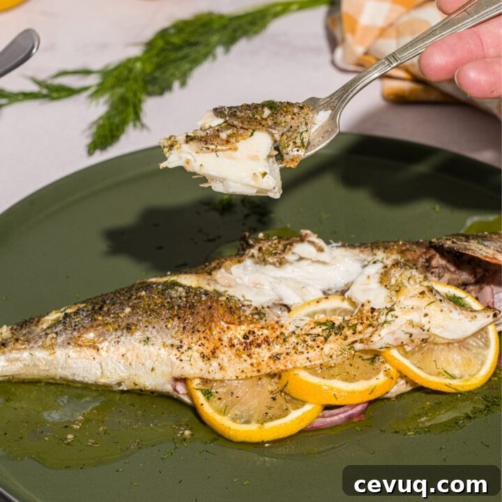 A green plate with greek whole roasted branzino stuffed with sliced lemon. There is a fork with a bite of branzino held over the plate. The plate is on a white counter with a yellow checkered napkin.
