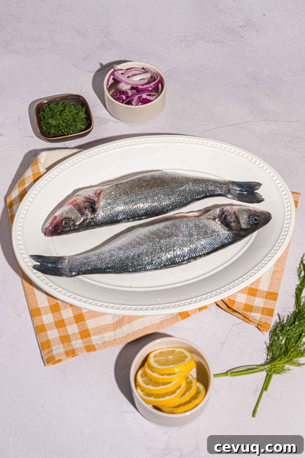 A white plate with two whole branzinos on a white counter with small bowls of lemon, herbs, and sliced red onion.
