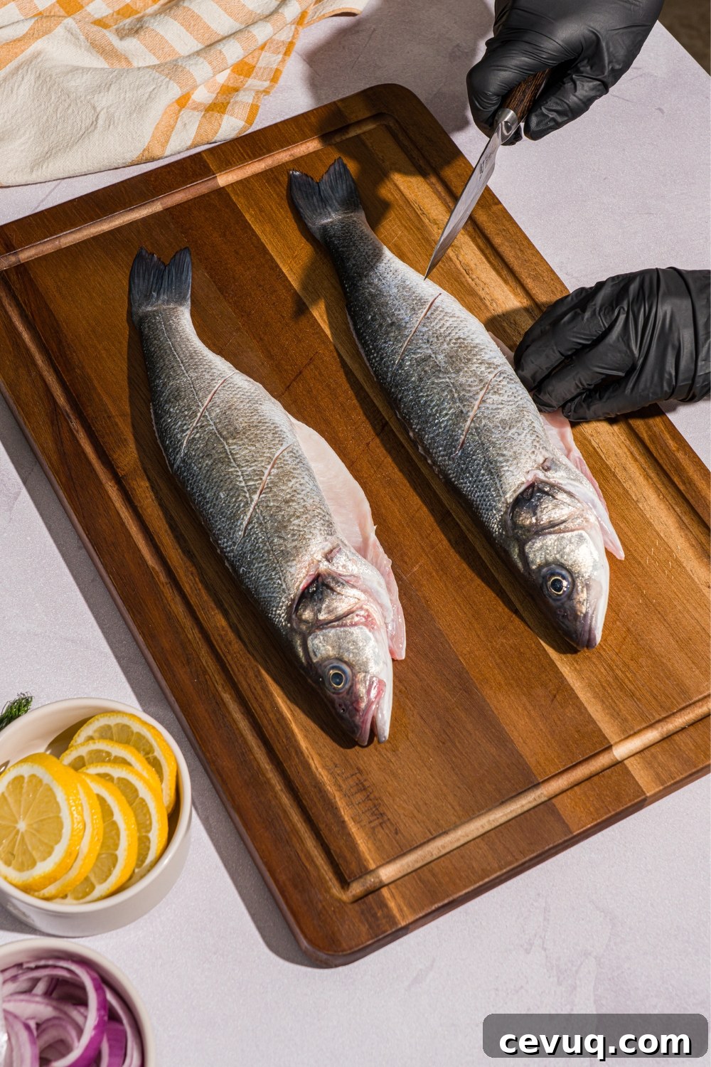 A wooden board with two whole branzinos. The branzinos are being slit with a knife. The board is on a white counter with a bowl of sliced lemons and yellow checkered napkin.