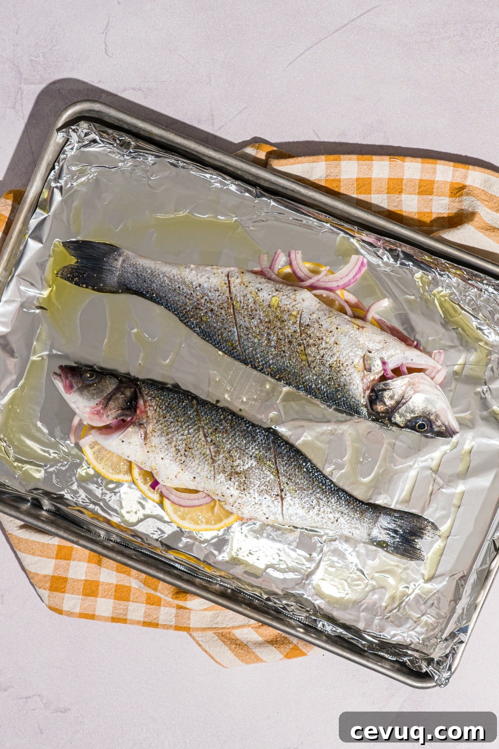 A baking sheet lined with foil with two whole branzinos stuffed with lemon and drizzled with oil. The sheet is on a white counter with a yellow checkered napkin.