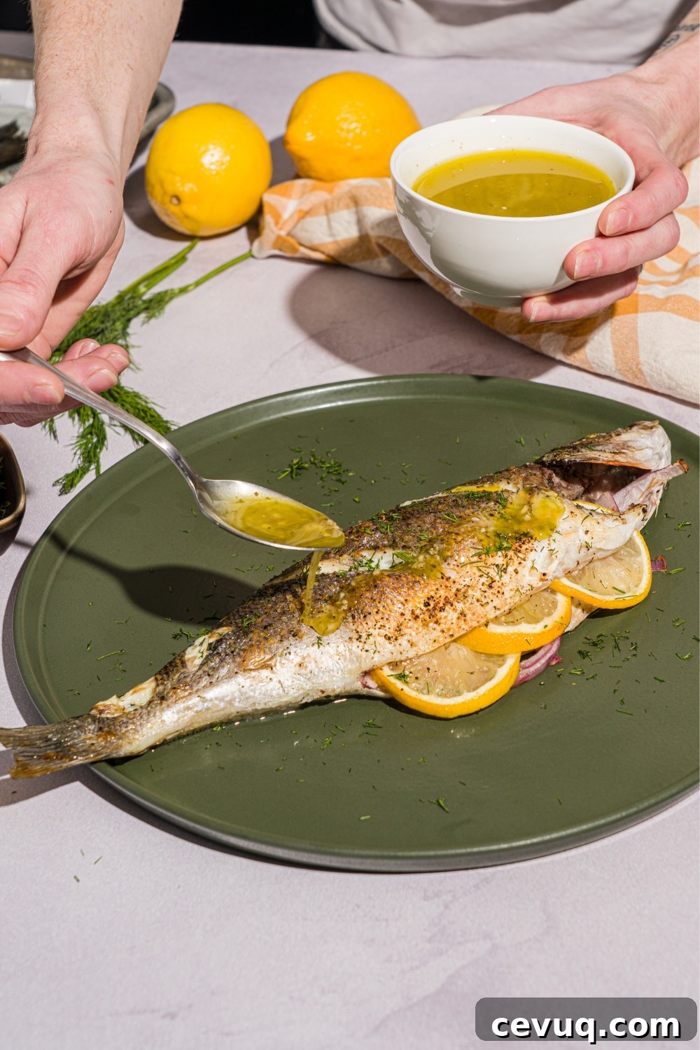 A green plate with greek whole roasted branzino stuffed with sliced lemon. A spoon is drizzling ladolemono dressing over the branzino. The plate is on a white counter with a yellow checkered napkin.