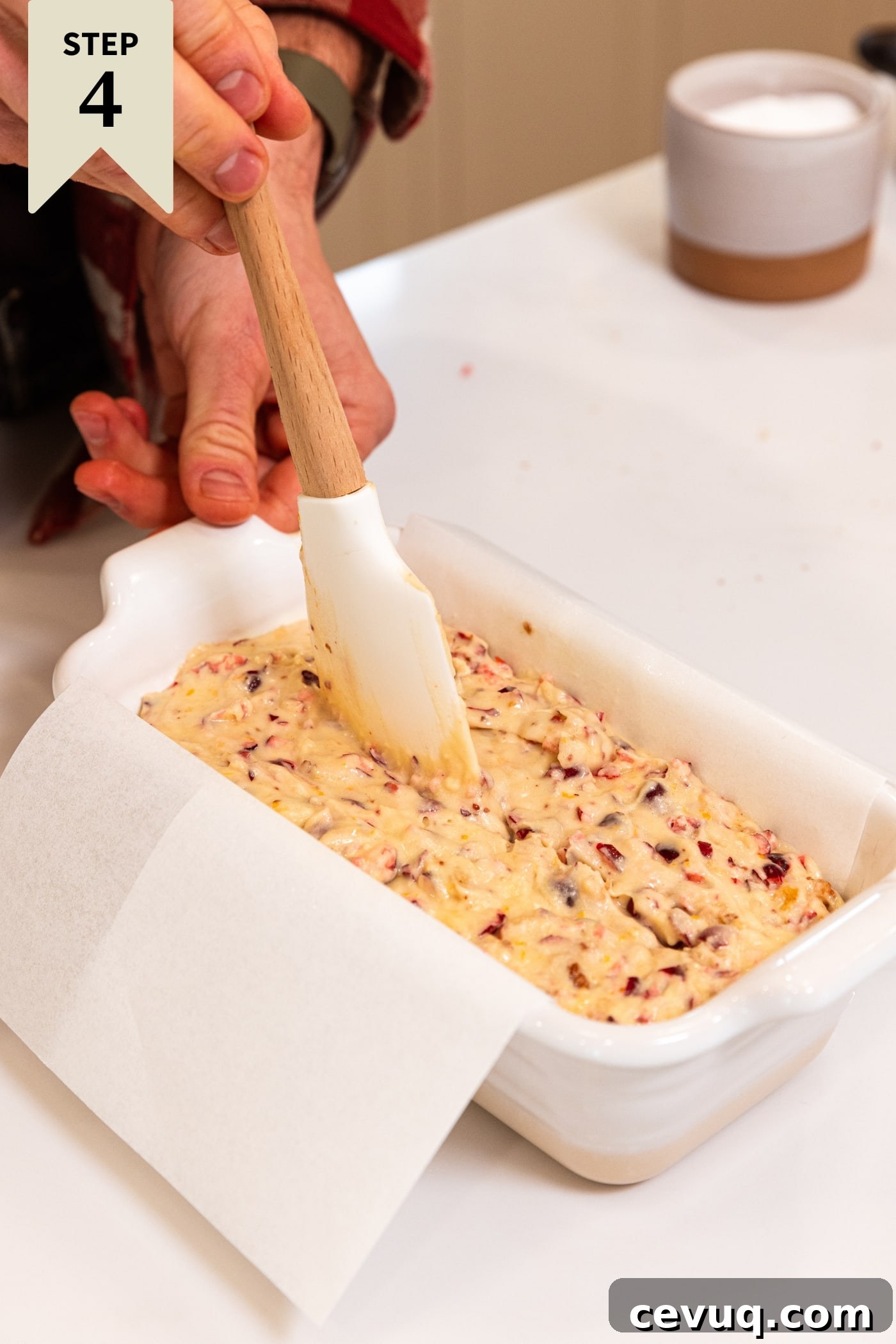 Hand pulling white spatula through the middle of cranberry orange bread dough in white loaf pan fitted with pieces of baking parchment all on white countertop.