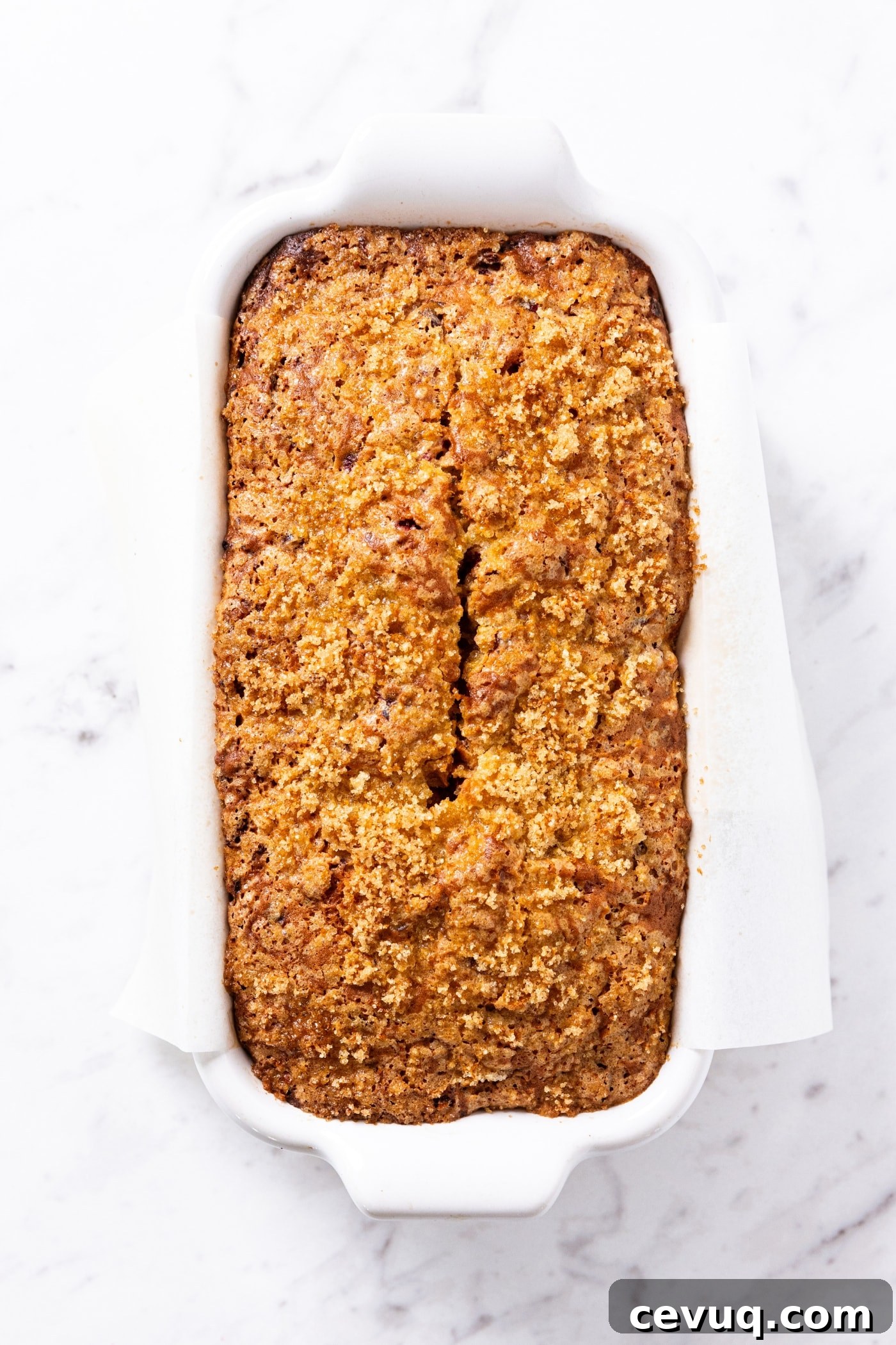 Baked orange cranberry bread sitting on white loaf pan on marble surface right after coming out of the oven.