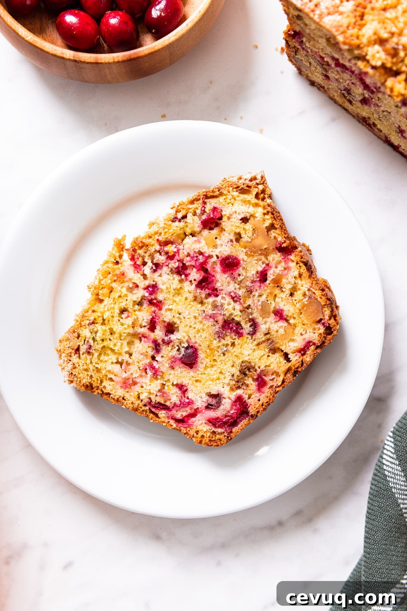 White marble surface with plate sitting with slice of cranberry orange bread with cranberries and rest of loaf of bread to the side.