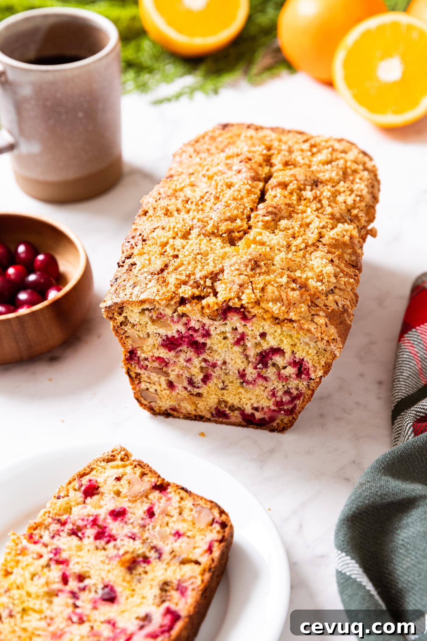 Loaf of cranberry orange bread sitting on white marble surface with slice of bread sitting on white plate with cup of coffee in the background along with oranges and cranberries.