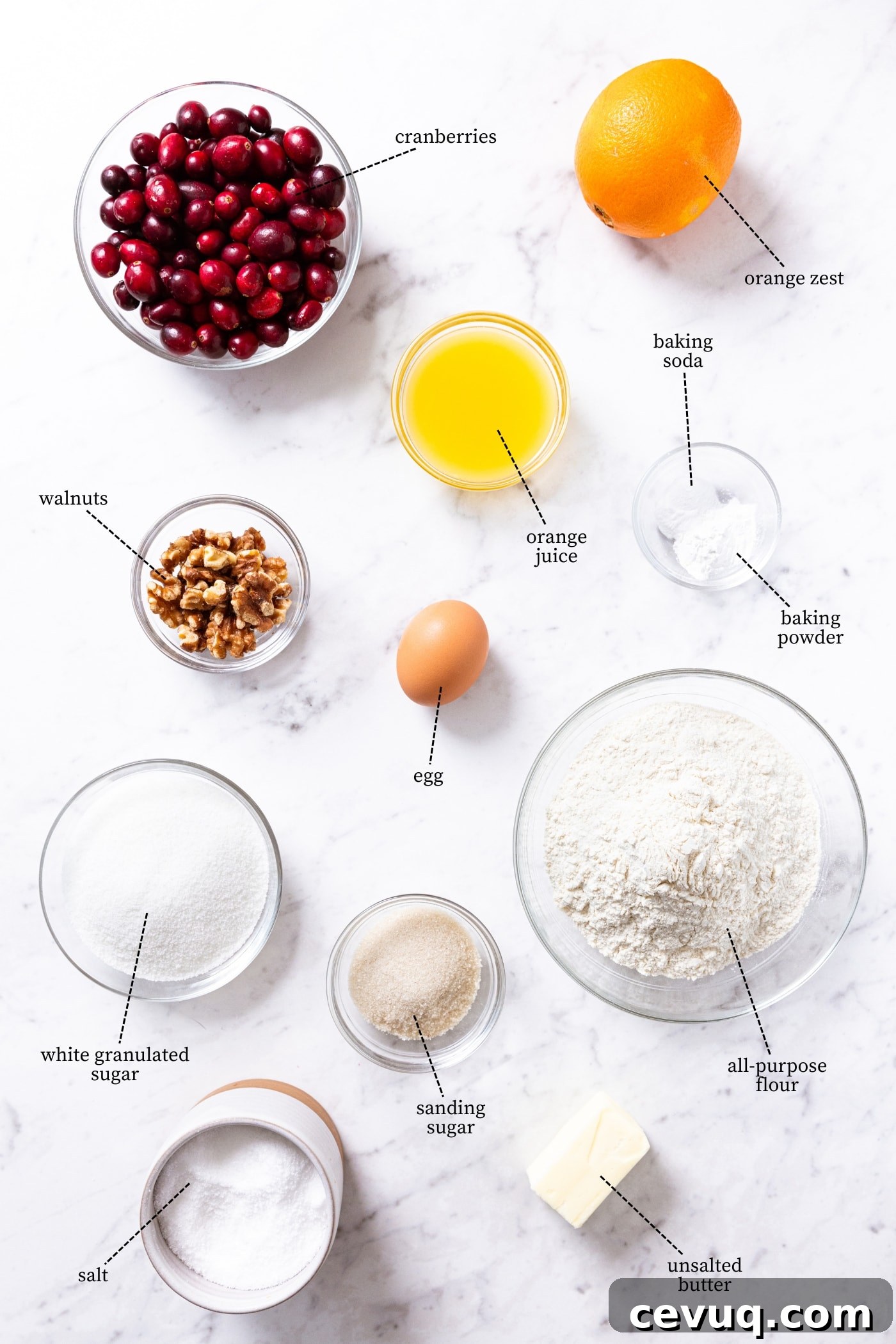 White marble surface filled with all ingredients needed to make cranberry orange bread including cranberries, orange juice, orange, flour, sugar, spices, and more.