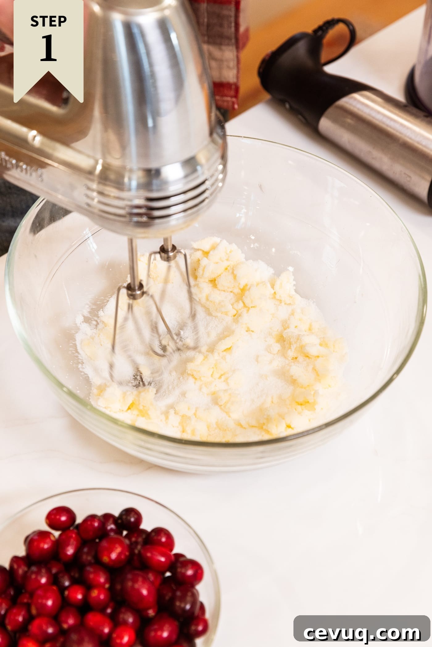 Glass mixing bowl with hand mixer spinning inside with butter and sugar being mixed together all on white surface with bowl of cranberries to the side.