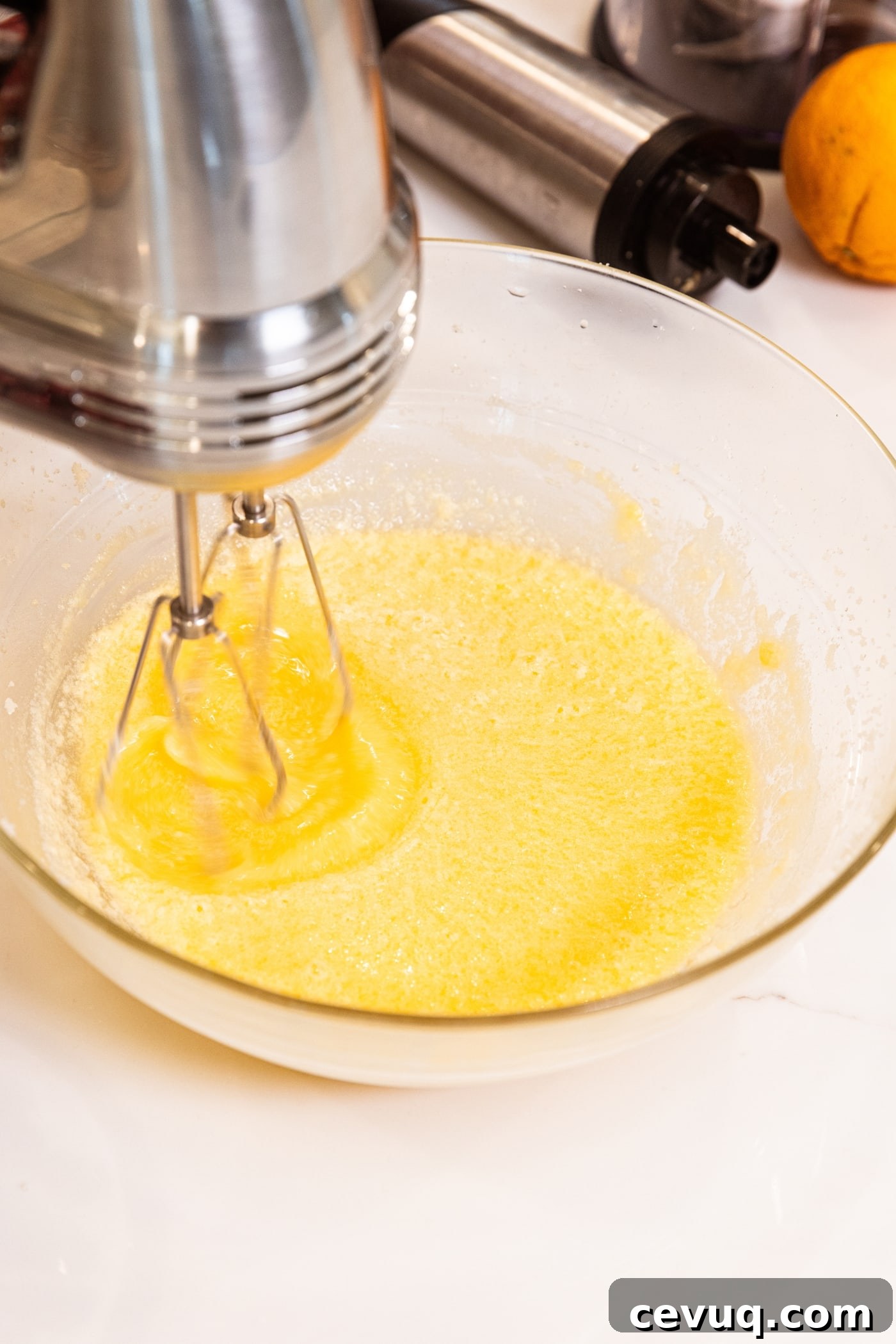 Hand mixer mixing together batter with light orange color on white surface with orange in the background.