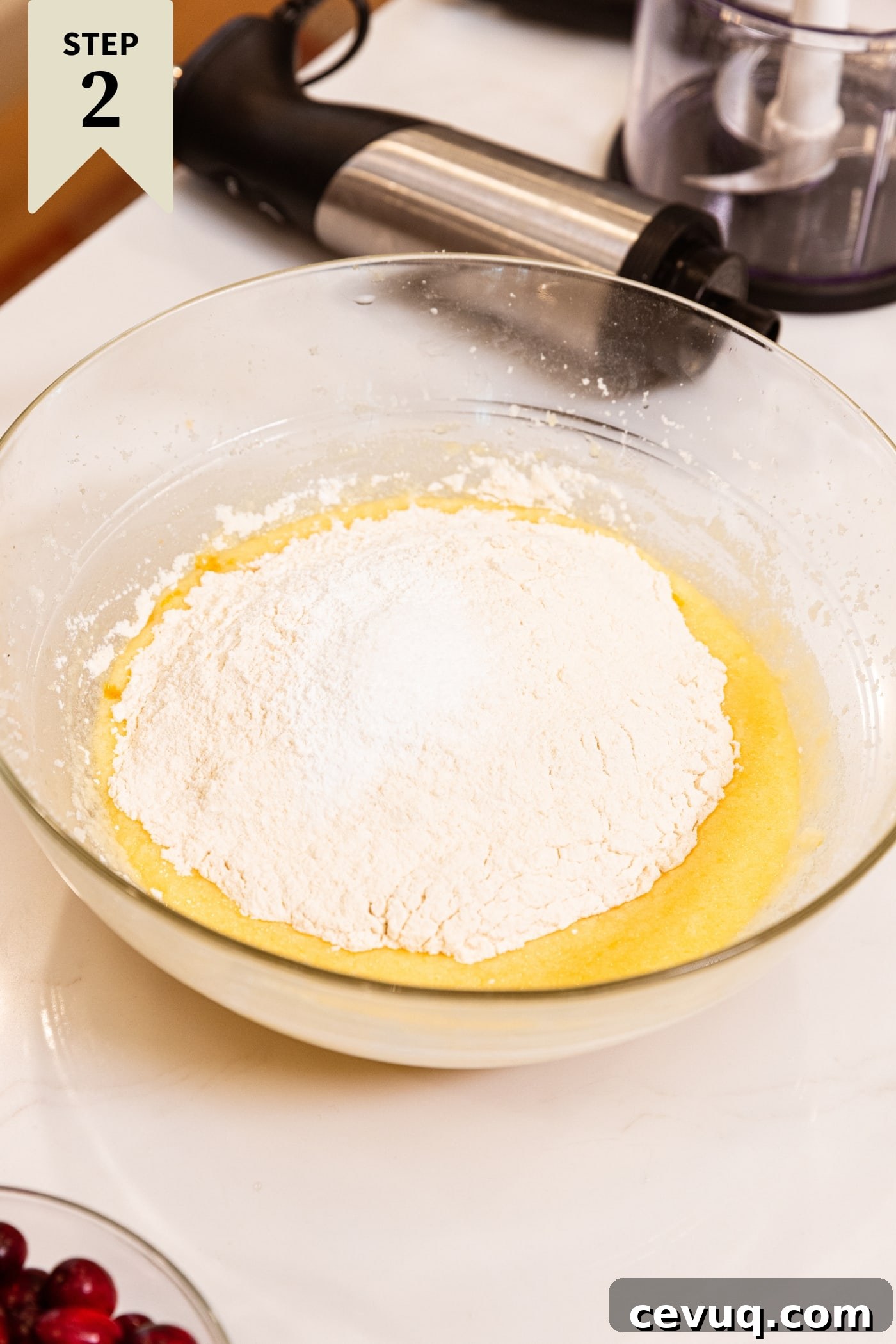 Glass mixing bowl filled with yellow colored batter with scoops of flour on top before being mixed with hand mixer all on white surface.