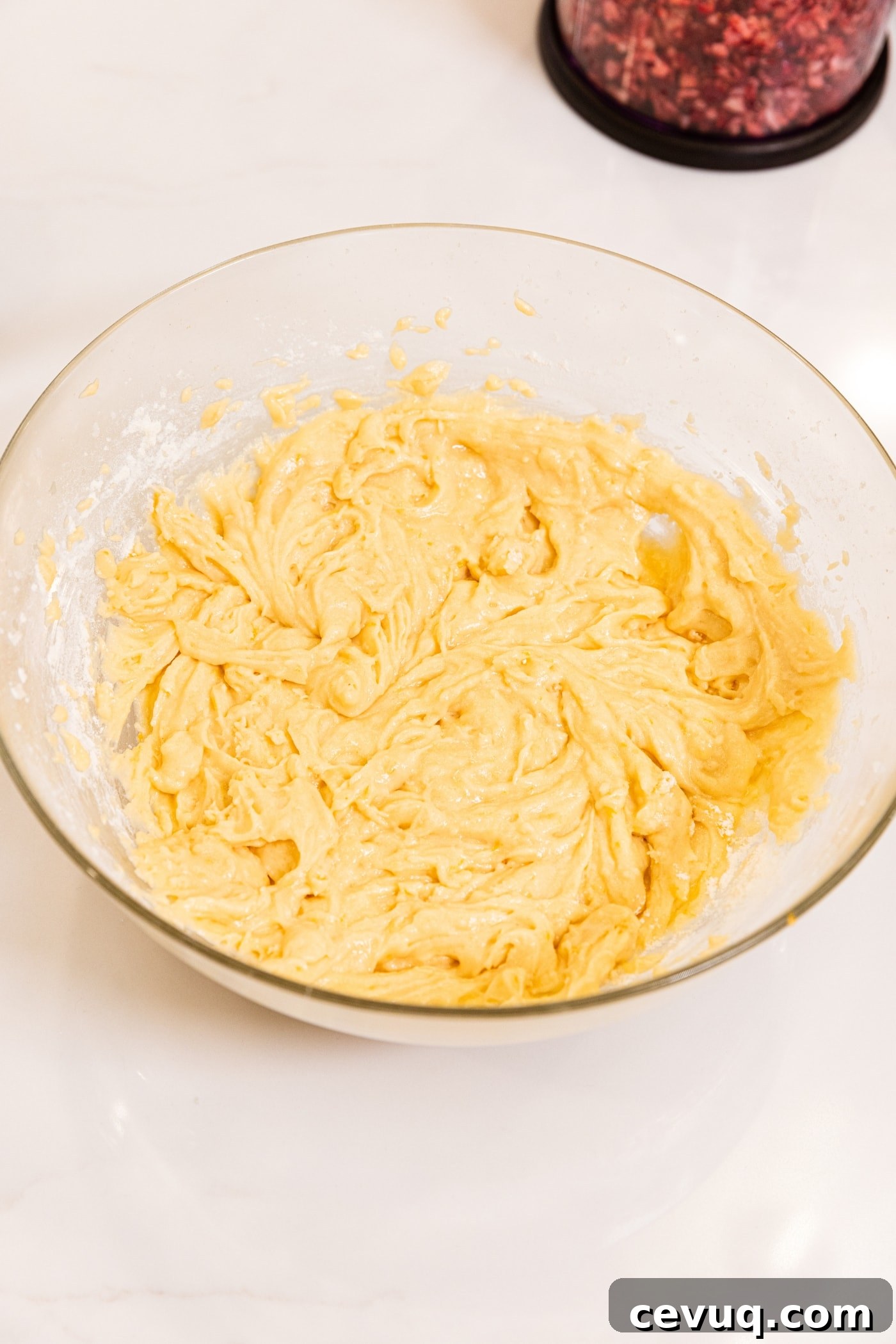 Glass bowl filled with light yellow colored batter after being mixed with a hand mixer sitting on white surface.