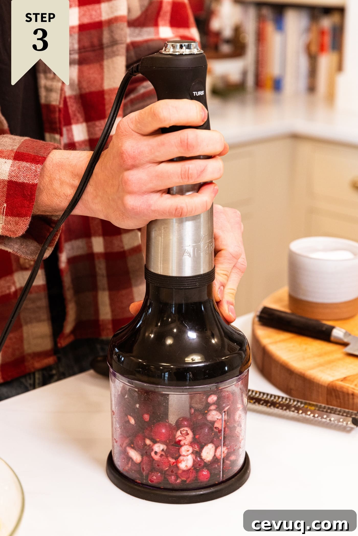 Hand holding food processor chopping up cranberries all on white surface with wood cutting board in background.