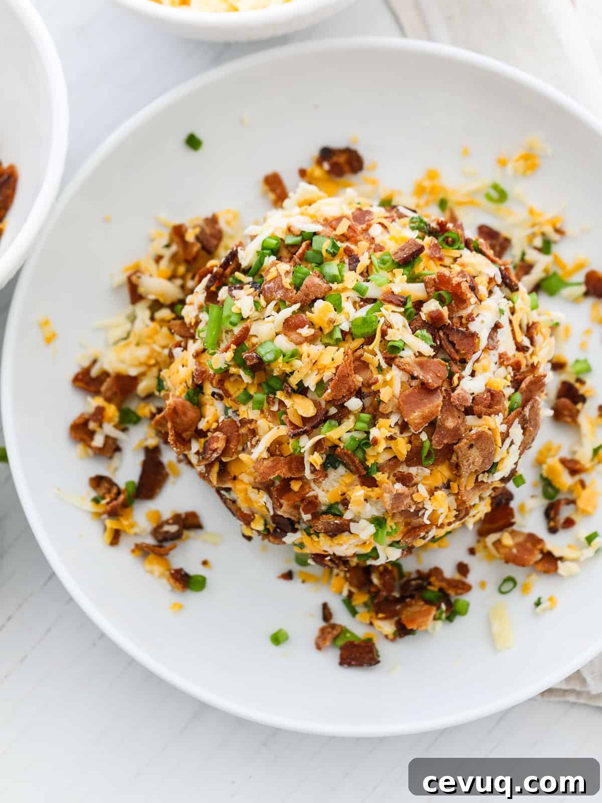 A bacon cheese ball coated on a plate with crumbled bacon, grated cheese, and green onions.