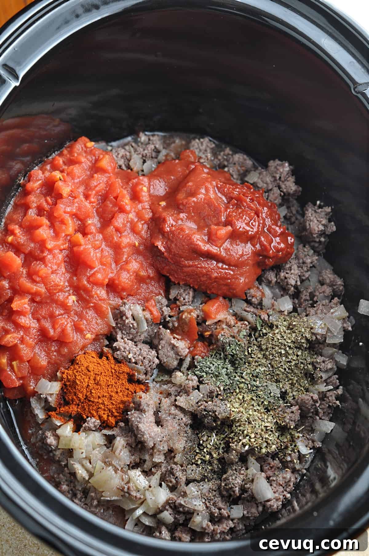 Tomatoes and seasonings on top of cooked ground beef and onions in slow cooker.