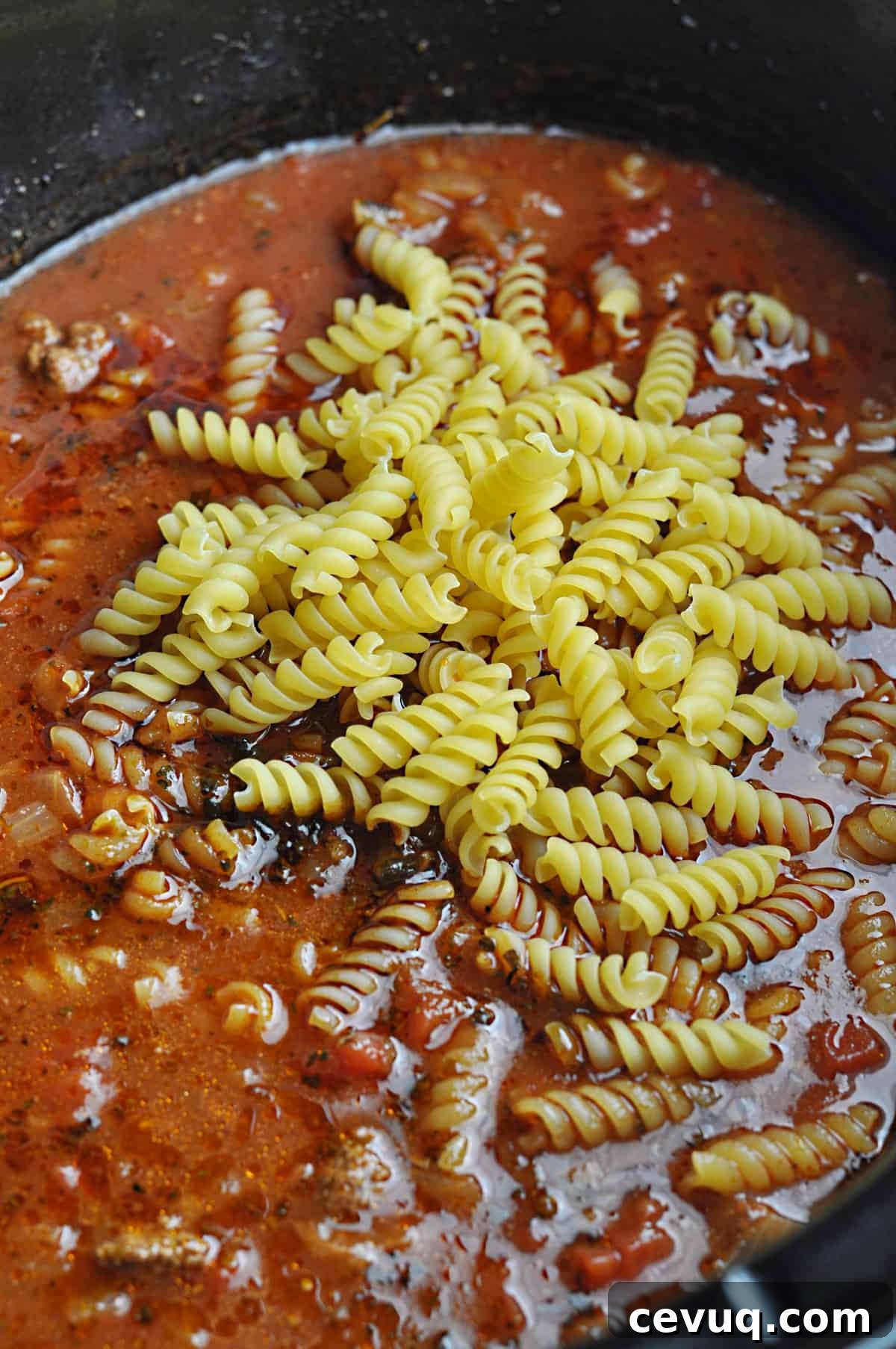 Uncooked pasta on top of ground beef and tomato mixture in slow cooker.