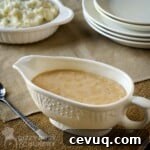 Side shot of gravy in a decorative ceramic dish with a tray of mashed potatoes and a wooden spoon in the background all on a burlap cloth.