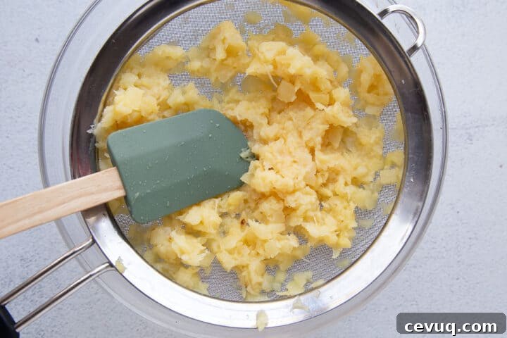 spatula pressing on crushed pineapple in a mesh colander.