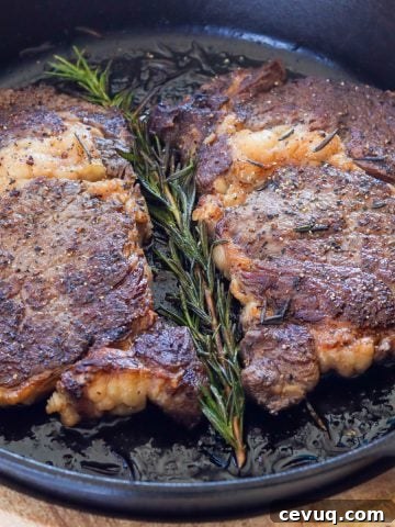 Two ribeye steaks in a cast-iron skillet with a sprig of rosemary.