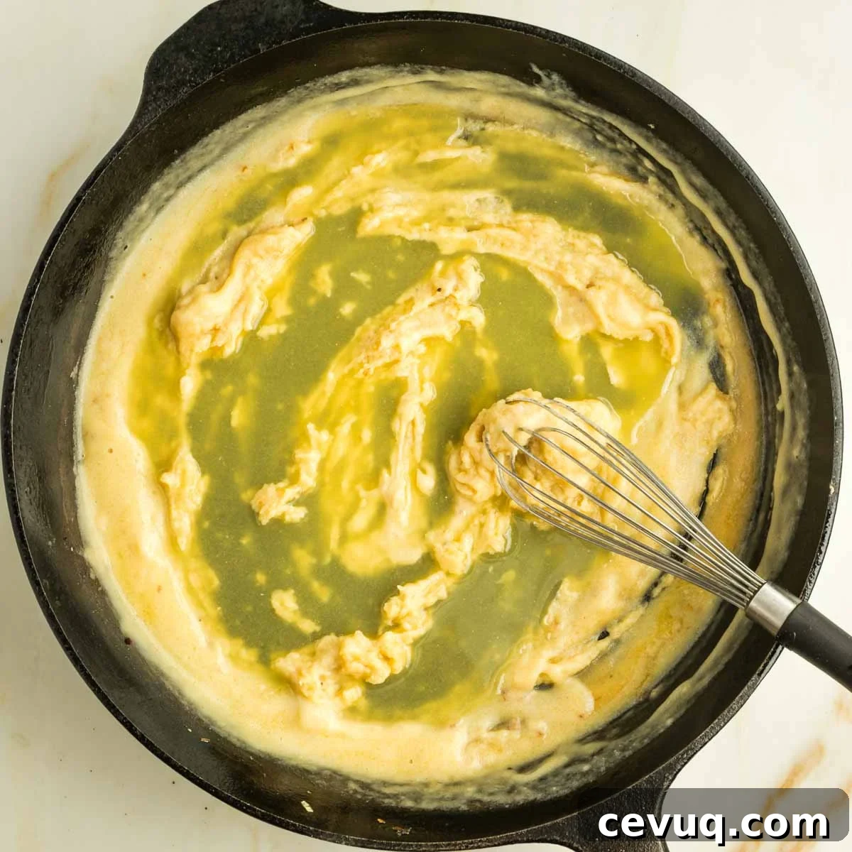 Broth poured onto a roux that's being stirred with a whisk in a cast iron skillet.